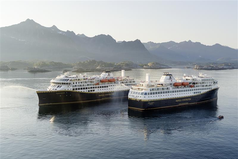 Zwei Postschiffe der Havila Kystruten (Havila Capella und Havila Castor) liegen nebeneinander in einem ruhigen Fjord vor einer dramatischen Bergkulisse in Norwegen.