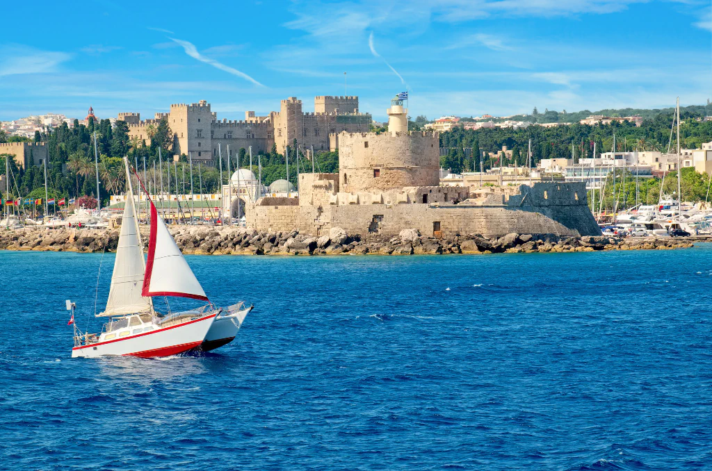 Ein Segelboot mit weißen Segeln und rotem Streifen auf blauem Meer im Vordergrund. Im Hintergrund eine historische Festung mit einem runden Turm und einer griechischen Flagge an einer felsigen Küste.
