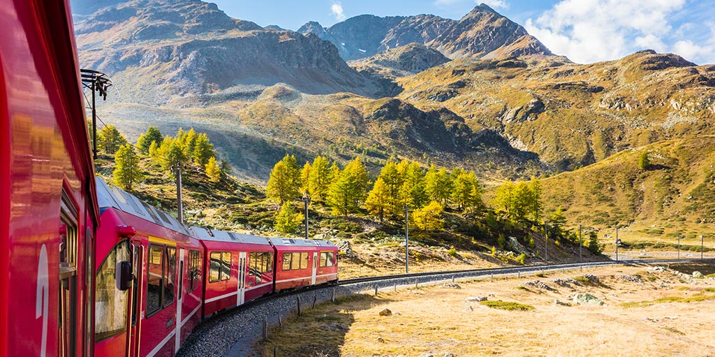 Ein roter Zug des Bernina Express fährt entlang einer malerischen Kurve durch eine alpine Landschaft. Die umliegenden Berge sind mit herbstlich gefärbten Bäumen und trockener, goldener Vegetation bedeckt. Der Himmel ist strahlend blau mit einigen weißen Wolken, was die Szene in warmem Licht erscheinen lässt. Ein roter Zug des Bernina Express fährt entlang einer malerischen Kurve durch eine alpine Landschaft. Die umliegenden Berge sind mit herbstlich gefärbten Bäumen und trockener, goldener Vegetation bedeckt. Der Himmel ist strahlend blau mit einigen weißen Wolken, was die Szene in warmem Licht erscheinen lässt.